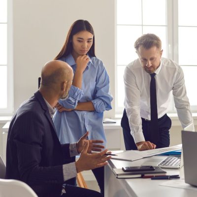 A group of business professionals strategizing about their workplace around a coworkers desk.
