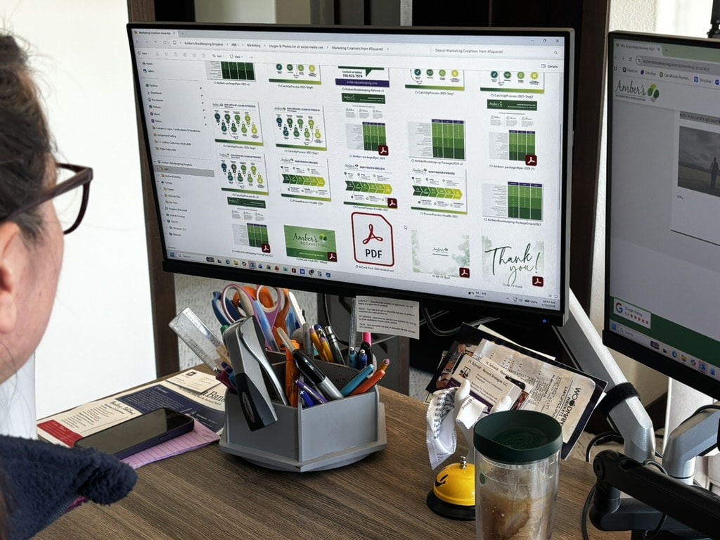 Bookkeeper reviewing client documents and marketing materials on a dual-monitor workstation