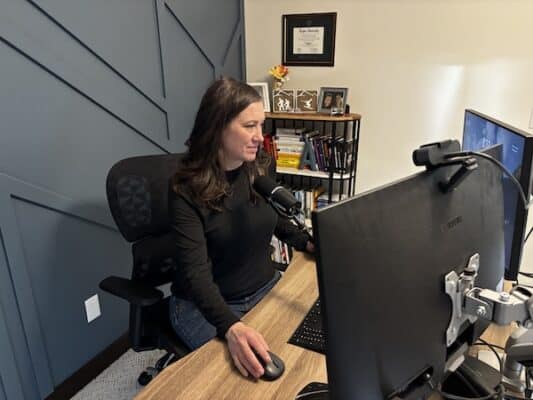 Amber Malone working at her desk in a modern home office, using dual monitors and speaking into a microphone while managing bookkeeping and accounting tasks.